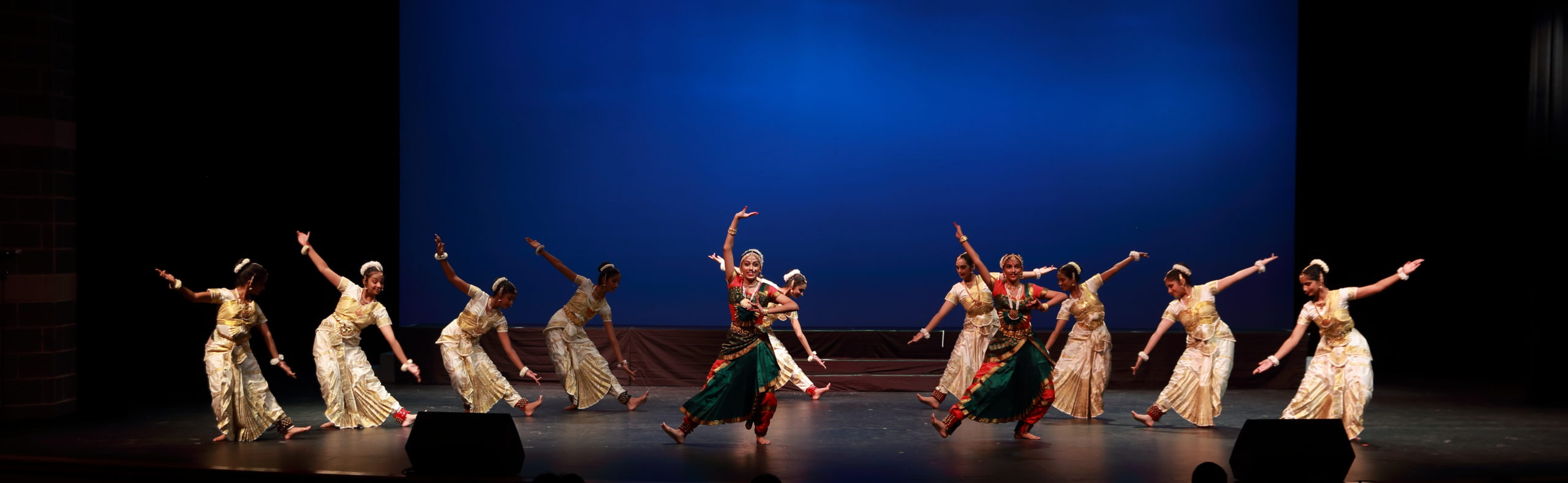 Ten dancers, 4 in white, 2 in green, and 4 in white, in formation on stage with a blue background